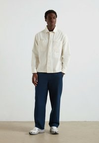 Uomo in piedi contro un muro bianco, indossa una camicia crema con bottoni, pantaloni blu navy e sneakers bianche, con una mano in tasca e l'altra rilassata sul fianco.