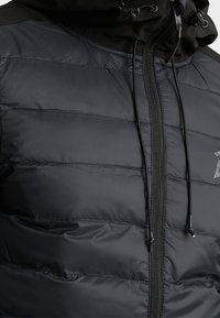 Svart kviltad jacka med framzipper och svarta justerbara dragsnören på huvan.