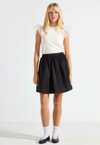 Top bianco con maniche a volant trasparenti, abbinato a una gonna nera fino al ginocchio. Indossato con ballerine nere e calzini bianchi alla caviglia. Tessuto liscio.