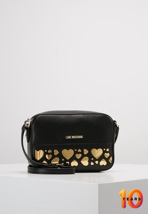 Schwarze Moschino Crossbody-Tasche mit goldenen Herz- Ausschnitten und verstellbarem Riemen, präsentiert auf einem weißen Podest.