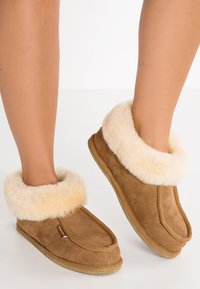 Pantofole in scamosciato marrone con polsino in pelliccia di pecora beige e soffice, punta arrotondata e suola in gomma strutturata. Materiale morbido e design accogliente.