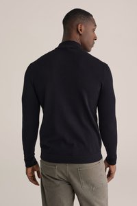 Tumši zils džemperis ar pieguļošu dizainu, garām piedurknēm un ribām pie manžetēm un apakšmalas. Modelis valkā olīvzaļas bikses, kas ir vaļīgas pie formas.