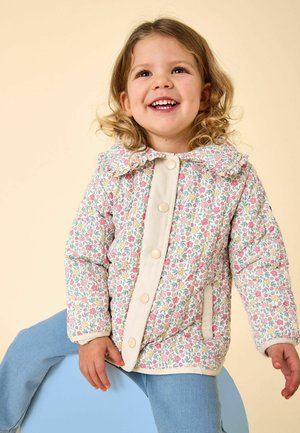 Copil mic cu păr creț zâmbind, purtând o jachetă cu model floral și nasturi mari, și pantaloni albaștri, așezat pe un obiect rotund albastru.