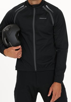 Homme portant une veste de cyclisme Endurance noire, tenant un casque de vélo Endurance noir sur un fond blanc.