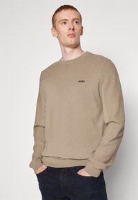 Beige knitted sweater with round neck, ribbed accents, and small black "BOSS" logo on the chest. Worn with dark jeans.