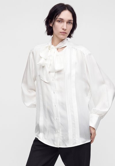 Jeune personne aux cheveux courts et foncés portant un chemisier blanc avec un grand nœud et un pantalon noir, debout devant un fond uni.
