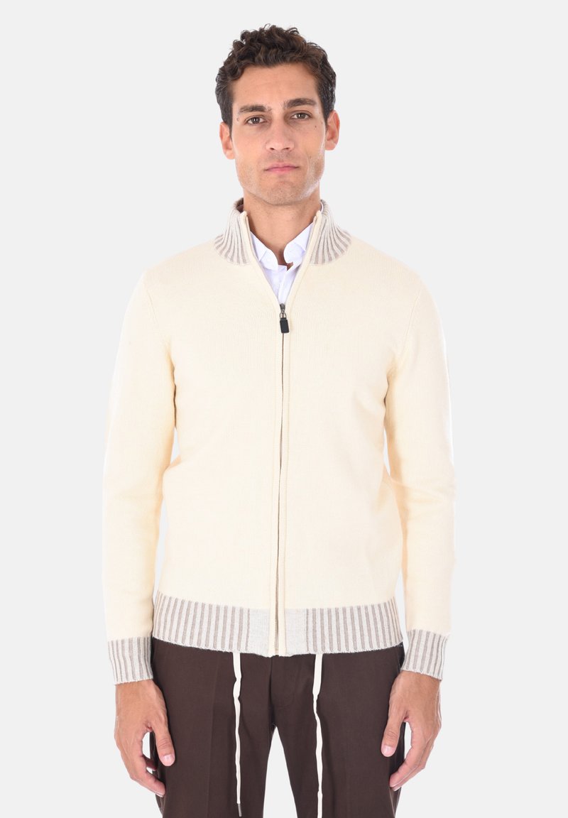 Maglione zip-up color crema con colletto e polsini a coste beige. Presenta un design slanciato e una vestibilità aderente. Indossato sopra una camicia bianca.