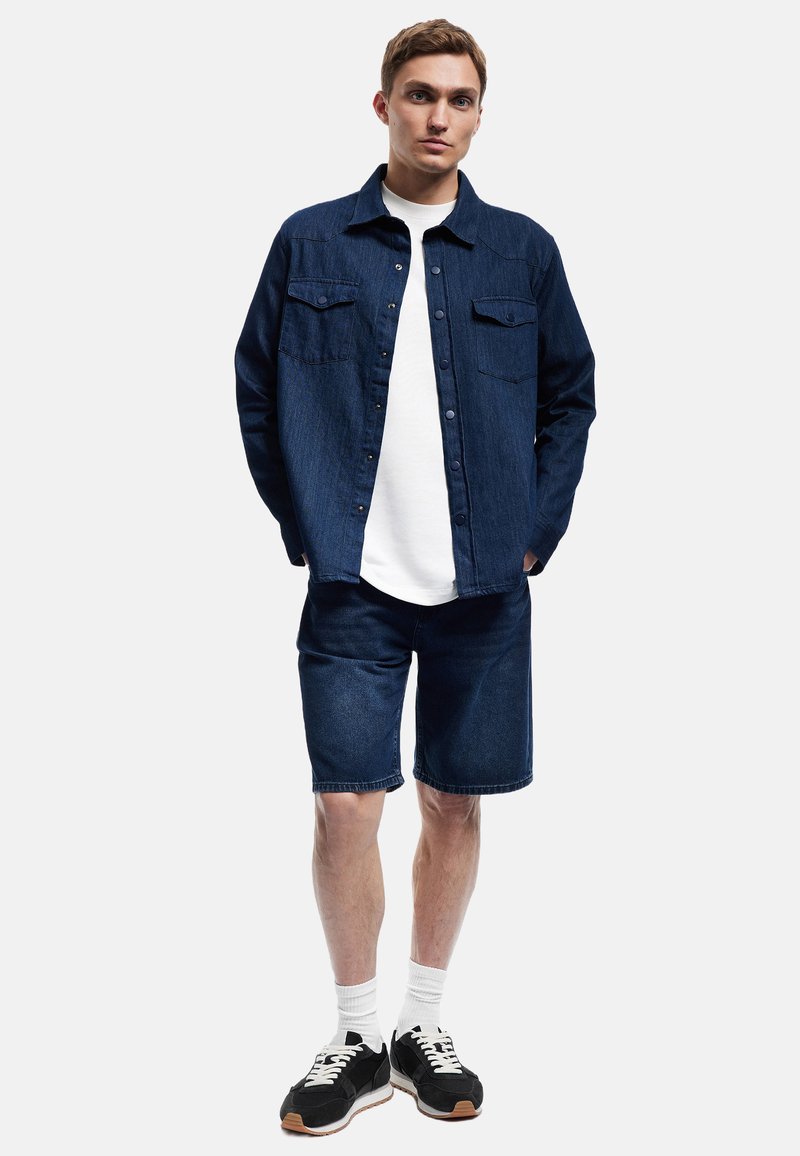 Camisa de denim en azul oscuro con dos bolsillos en el pecho, combinada con shorts de denim azul oscuro a juego. Zapatillas negras con acentos blancos completan el atuendo.