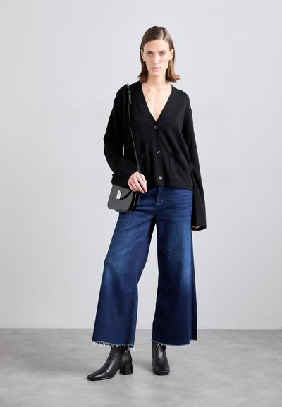 Cardigan preto com um decote em V profundo, duas bolsos frontais, combinado com jeans de perna larga azul escuro com bainhas desfalcadas e botas negras de tornozelo.