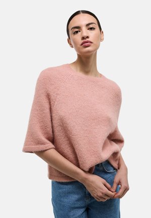 Femme aux cheveux foncés portant un pull à manches courtes rose et un jean bleu, debout devant un fond clair uni.