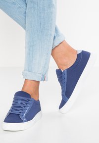 Baskets tricotées bleues avec un accent de talon bleu lisse, semelle en caoutchouc blanche, et lacets assortis. Jean en denim roulé partiellement visible.