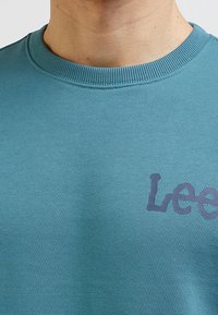 Sudadera en color azul verdoso con un cuello redondo acanalado y un logotipo texturizado de "Lee" en un tono azul más oscuro en la parte delantera. La tela parece suave y lisa.