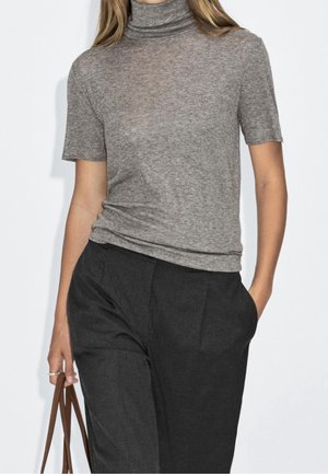 Persoon draagt een grijze korte mouw coltrui, gestopt in zwarte geplooide broek, houdt bruine tasgrepen vast in de linkerhand.