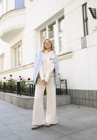 Lichtblauwe cardigan, crèmekleurige hoodie met "BIZU" logo, crèmekleurige brede broek en beige schoenen, tegen een achtergrond van een wit gebouw en rozenstruiken.