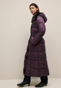 Lang lilla pufferjakke med hætte, quiltet design, elastisk talje og sidelommer, parret med sorte hælede ankelstøvler.