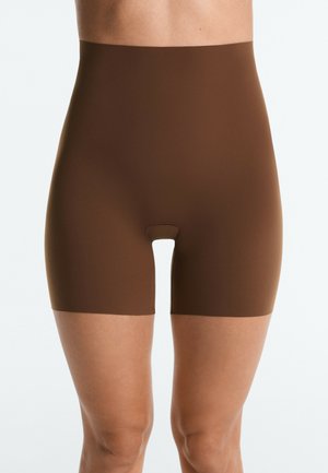Prim-plan cu o persoană care poartă pantaloni scurți modelatori maro, fără cusături, cu talie înaltă, care se întind până la jumătatea coapsei, pe un fundal simplu.