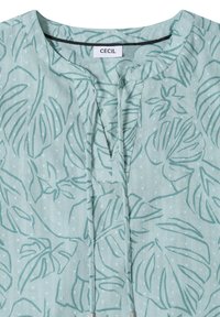 Blouse vert clair avec de longues attaches, une texture subtilement pointillée et un motif de feuilles tropicales vert foncé, avec un col rond.