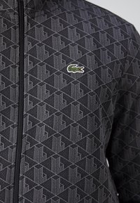 Veste noire zippée avec motif géométrique texturé et petit logo crocodile vert sur la poitrine.