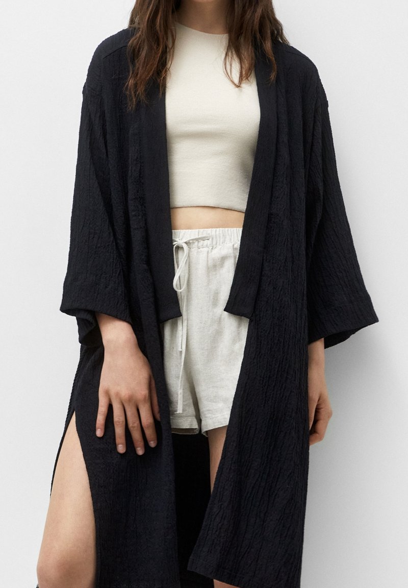 Ropa estilo kimono negra con un acabado texturizado, con abertura frontal y mangas anchas. Lleva puesta una blusa corta de color gris claro y pantalones cortos blancos.