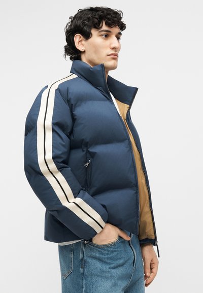 Mees seljas sinine puffer-jakk, mille varrukatel on valged ja mustad triibud, beež sisevooder paistmas, ja sinised teksad, seisab parema käe taskus.