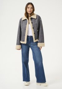 Grijze neppelschoenen jas met beige bies en grote knopen, gecombineerd met uitlopende blauwe jeans en witte sneakers. De stof heeft een zachte textuur.