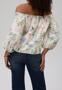 Blusa off-the-shoulder in tessuto crema con motivo floreale che presenta fiori rosa e viola, maniche a sbuffo e scollatura arricciata.