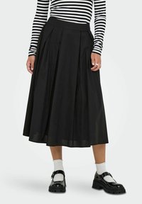 Gonna nera plissettata con vita aderente, realizzata in tessuto leggero. Abbinata a scarpe nere e calzini bianchi alla caviglia.