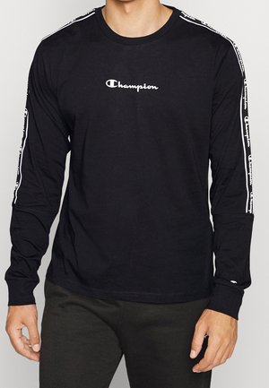 Mann trägt schwarzen, langärmligen Champion-Shirt mit weißem Logo auf der Brust und markierten Streifen an beiden Ärmeln.