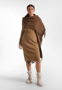 Braunes geripptes Kleid mit passendem braunen Poncho, der Fransenränder aufweist. Kombiniert mit beigen Schnürstiefeln. Glatte Textur über das gesamte Material.