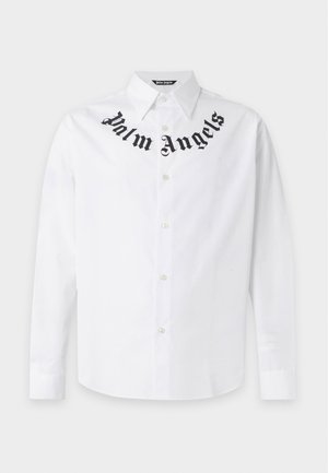 Mann trägt ein weißes Langarmshirt mit schwarzer Schrift am Kragen, braune kniehohe Shorts und schwarze Knöchelstiefel, steht mit den Händen in den Taschen.