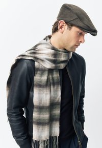 Gray and cream plaid scarf with frayed edges draped over a black leather jacket. The model wears a brown flat cap.