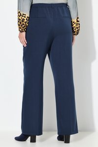 Vrouw draagt marineblauwe wijde broek met elastische tailleband, gecombineerd met zwarte enkellaarsjes met hak en een grijze blouse met luipaardprint mouwen.