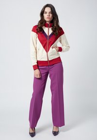 Giacca a zip crema con motivi geometrici rossi e viola; abbinata a pantaloni larghi viola e scarpe con tacco viola.