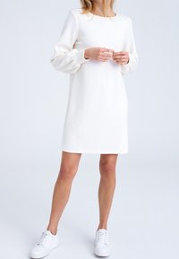 Vit klänning med långa, puffiga ärmar, rundad halsringning och rak silhuett. Kombinerad med vita sneakers för en avslappnad look.