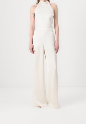 Vrouw draagt een mouwloze witte halter jumpsuit met wijde broekspijpen, staand tegen een effen witte achtergrond.
