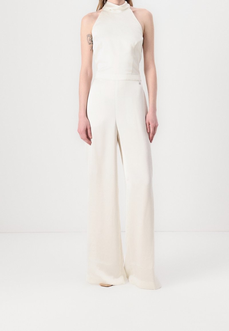 Femme portant une combi-pantalon blanche sans manches à col halter avec un pantalon à jambes larges, debout devant un fond blanc uni.