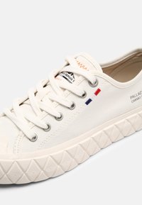Baskets en toile blanches avec semelle texturée, lacets blancs, œillets métalliques et petit détail de drapeau rouge, blanc et bleu brodé sur le côté.