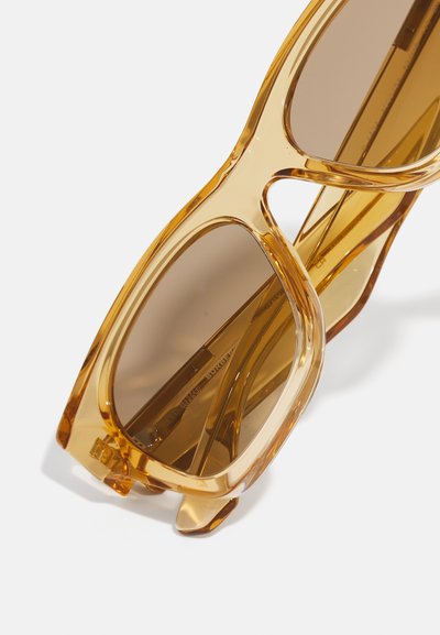Translucente bernsteinfarbene Sonnenbrille mit braun getönten Gläsern, breiten rechteckigen Rahmen und einer glatten Kunststoffoberfläche. Bemerkenswerte Designelemente sind dünne Akzente.