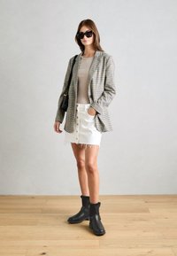 Blazer à carreaux, haut beige, mini jupe blanche avec des boutons et des franges, bottines noires avec finition en fourrure et sac à main noir. Fond en parquet.