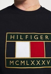 Detailní záběr na černou košili s vyšívaným logem Tommy Hilfiger, které obsahuje zelené, bílé, červené a námořnické bloky a římské číslice MCMLXXXV.