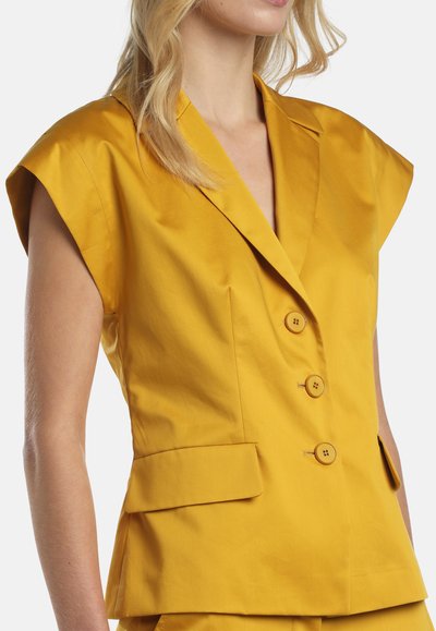 Nő sárga mustárszínű, ujjatlan blézert visel, amelynek strukturált válltömései, hajtókás gallérja, három gombja és lenyíló zsebei vannak.