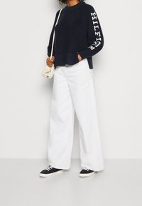 Marineblauer gerippter Pullover mit weißen Logodetails an den Ärmeln, kombiniert mit lockeren weißen Hosen und marineblauen Sneakers; minimalistischer Umhängebeutel.