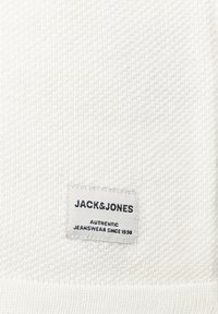Valge kudumine, millel on õmmeldud silt, millel on tekst "JACK&JONES AUTHENTIC JEANSWEAR SINCE 1990." Sile tekstuur ja minimaalne detailide arv.