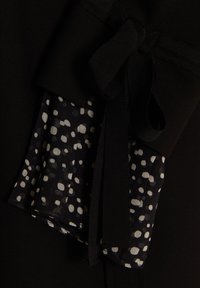 Robe noire avec un tissu texturé recouvert d'un motif à pois en blanc et gris. Fini avec un nœud en satin noir.