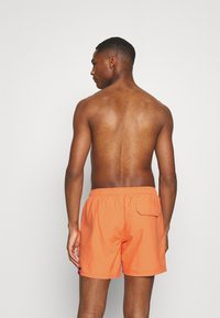 Oransje svømmeshorts laget av lett stoff, med elastisk midje, sidelomme og en logo på venstre ben.