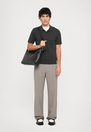 Borsa a spalla nera in pelle, polo grigia scura, pantaloni grigi chiari su misura, sandali neri con suole bianche, persona in piedi su uno sfondo bianco.