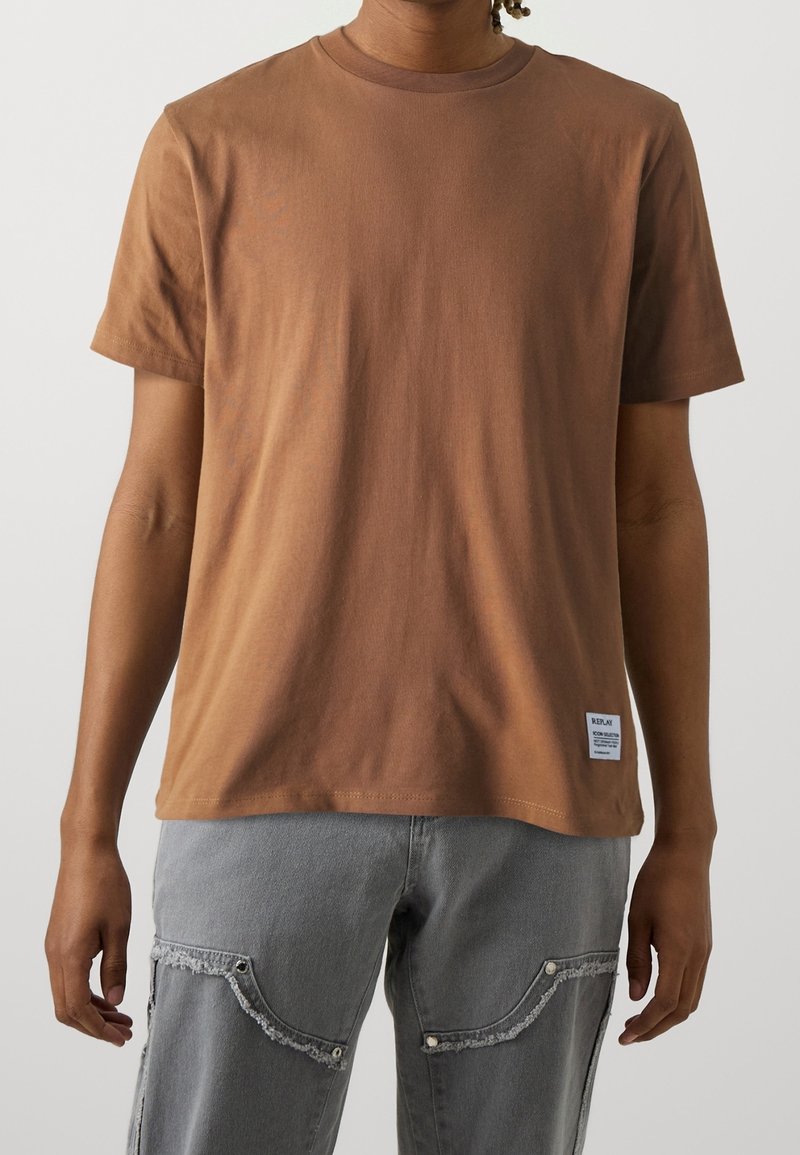 T-shirt en coton marron avec col rond, manches courtes et une étiquette en bas à gauche. Texture lisse, coupe décontractée, design minimaliste.