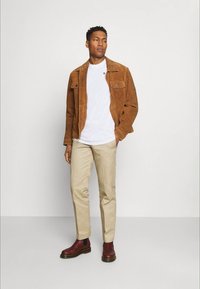 Brun mockaskinnjacka, vit t-shirt, beige byxor och mörkröda ankelstövlar bärs av en stående manlig modell mot en enkel bakgrund.