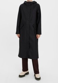 Černý voděodolný dlouhý kabát s předním zapínáním na knoflíky, kapucí a dvěma bočními kapsami; vyrobený z hladkého materiálu; spojený s hnědými kalhotami a světlými botami.