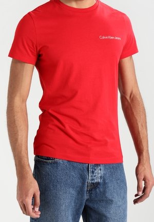 Homme portant un T-shirt rouge ajusté Calvin Klein Jeans et un jean en denim bleu, debout les bras détendus le long du corps.